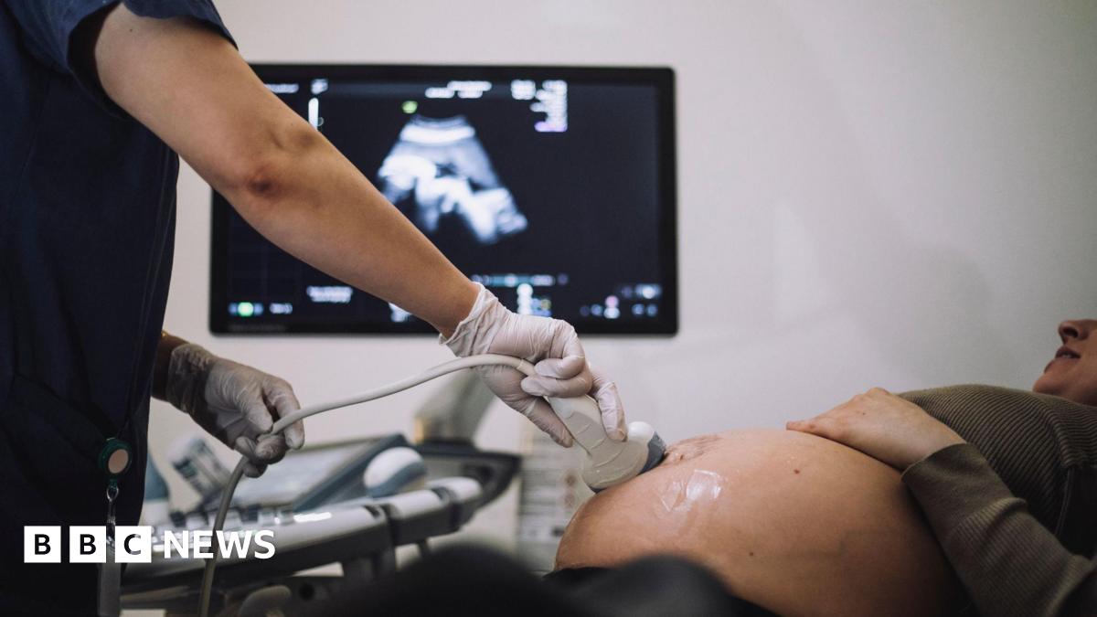 Stock shot of a pregnant woman being given an ultrasound scan. You can see the lady's pregnant belly and a scanning tool being operated by a medical practitioner wearing gloves. Behind is an out-of-focus image of the baby appearing on screen.