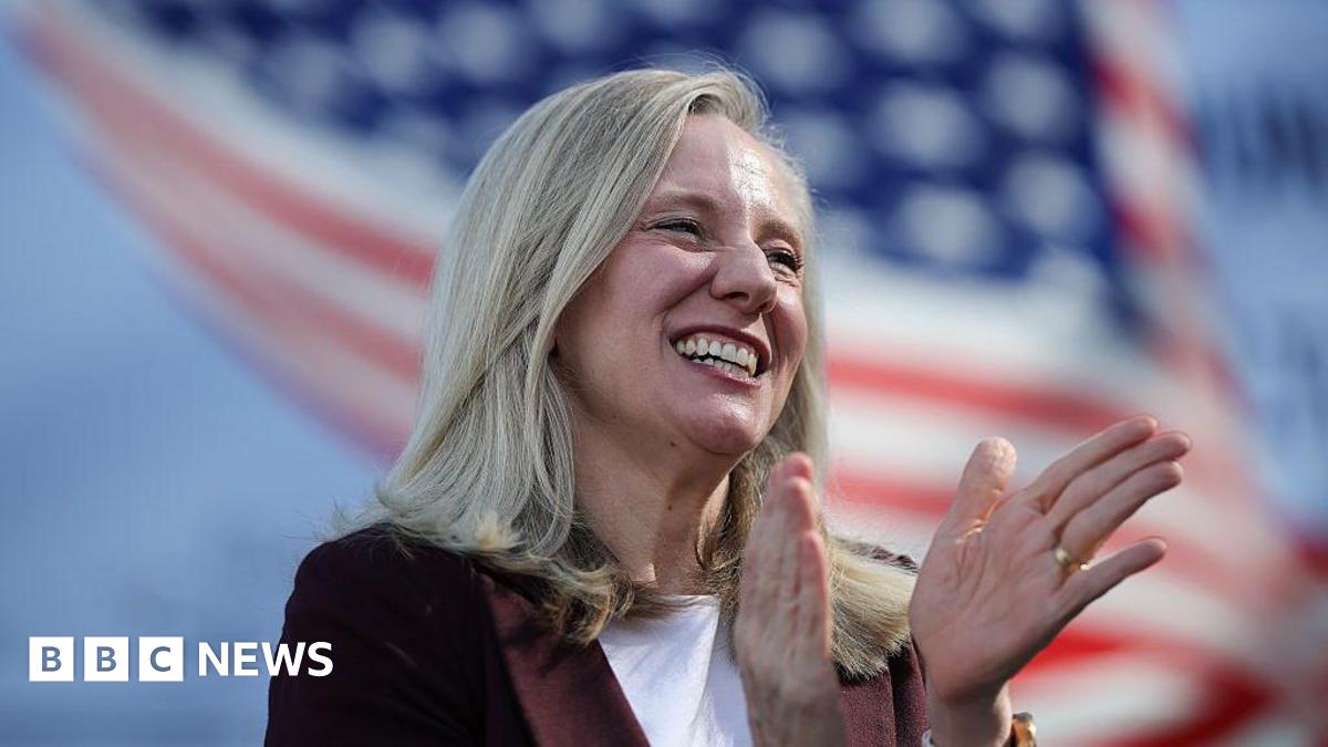 Spanberger smiles and holds out a hand. There's a US flag in the background