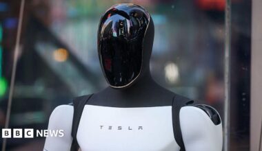 A Tesla Optimus robot. It is shaped like a human. We can see its head and shoulders. It has a sleek black reflective head, and a white torso and arms. In the middle of the torso is the word Tesla.