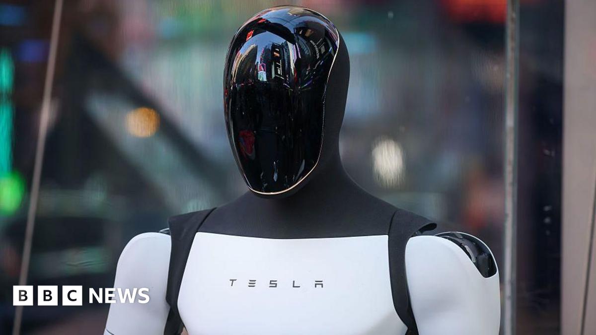 A Tesla Optimus robot. It is shaped like a human. We can see its head and shoulders. It has a sleek black reflective head, and a white torso and arms. In the middle of the torso is the word Tesla.