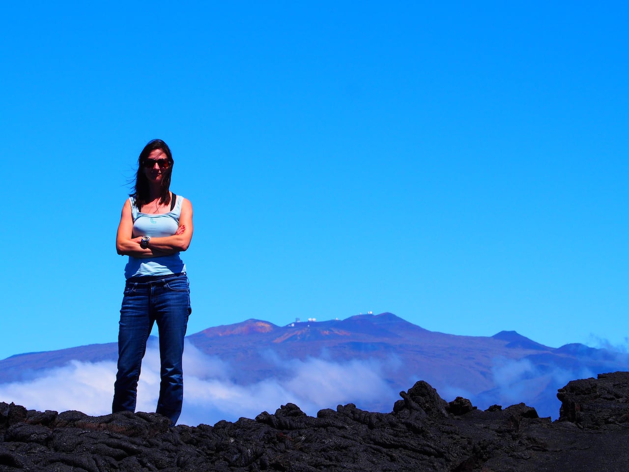 A woman with crossed arms stands on a mountain with a blue sky in the background.