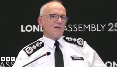 Sir Mark Rowley is seen sitting at a table speaking into a microphone, with a London Assembly backdrop behind him.