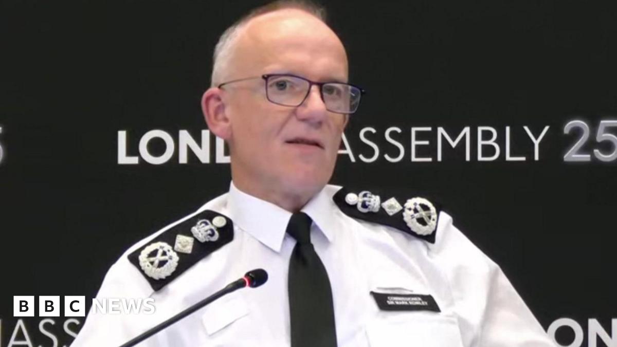 Sir Mark Rowley is seen sitting at a table speaking into a microphone, with a London Assembly backdrop behind him.