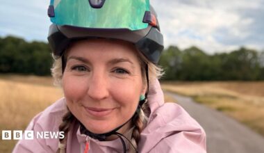 Claire Sharpe wears a cycling helmet and pink jacket has taken a selfie whilst riding her bike through a rural area. She has blonde hair and is smiling.