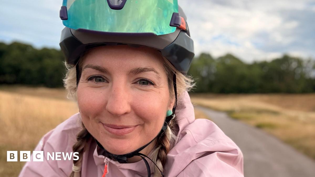 Claire Sharpe wears a cycling helmet and pink jacket has taken a selfie whilst riding her bike through a rural area. She has blonde hair and is smiling.