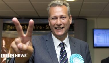 A grey-heaired man with a grey beard wearing a grey coat, blue shirt and a blue tie looking at the camera while walking away from the Old Bailey