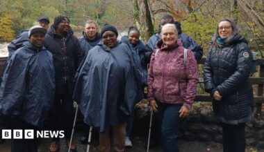 A mixed group of people stand in front of a fence by a river, posing for a picture. They all wear waterproof coats and winter clothing.