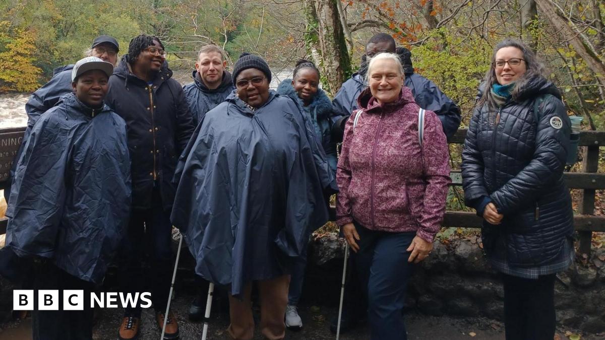 A mixed group of people stand in front of a fence by a river, posing for a picture. They all wear waterproof coats and winter clothing.