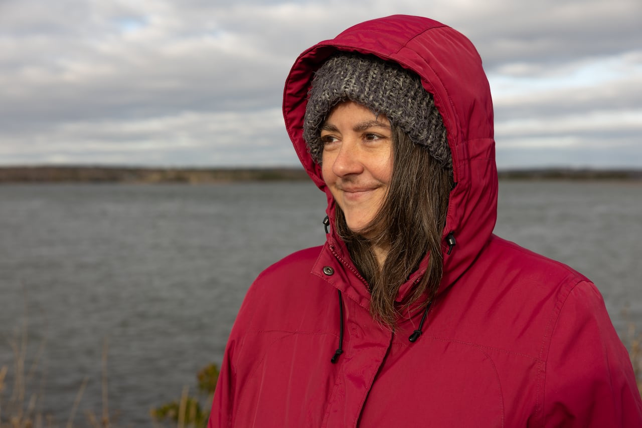 A woman in a red coat and grey tuque smiles. Behind her, water and trees are seen in the distance,