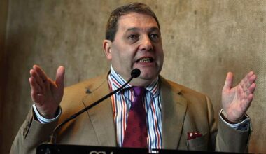 A brown-haired whire man wearing a brown suit, stripy shirt and claret tie stands in front of a microphone with his hands out gesturing