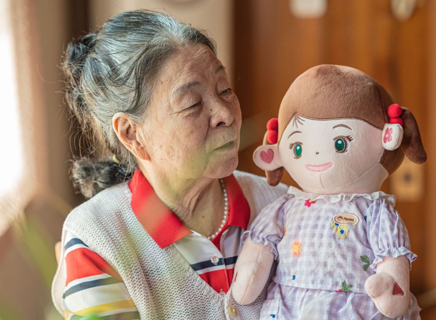 Elderly woman engages with an AI-powered Hyodol plushie robot companion. The doll is designed to help with daily care management by offering reminders to take medication or keep appointments through verbal interactions.