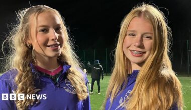 Two young girls are smiling for the camera. They are both wearing purple zip up jumpers with their football club branding on them in white. They are standing on a football pitch which is lit up with flood lights. They both have long blonde hair and braces.