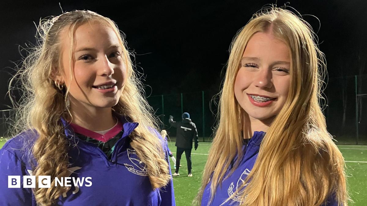 Two young girls are smiling for the camera. They are both wearing purple zip up jumpers with their football club branding on them in white. They are standing on a football pitch which is lit up with flood lights. They both have long blonde hair and braces.