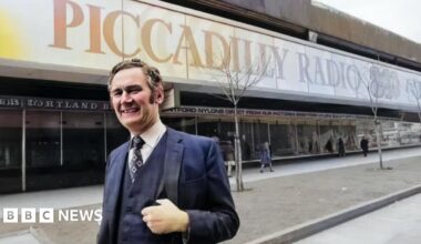 Philip Birch stands outside Piccadilly Radio's studios. He's wearing a suit including waistcoat. The Piccadilly Radio 261 text and logo adorns the outside window behind him.