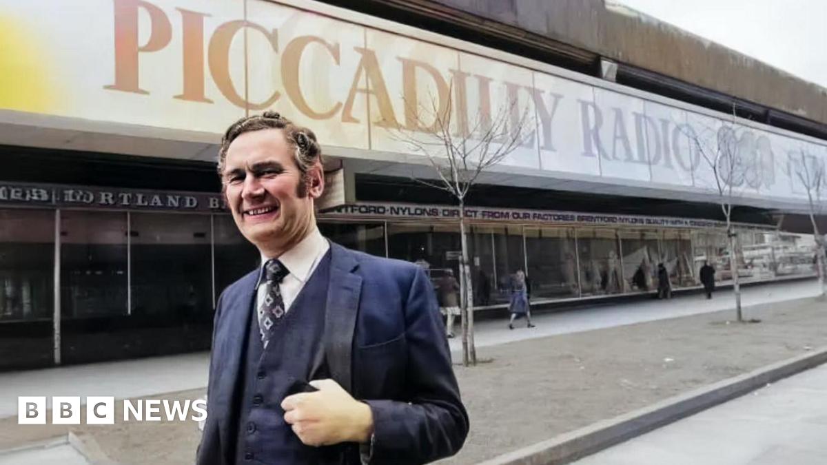 Philip Birch stands outside Piccadilly Radio's studios. He's wearing a suit including waistcoat. The Piccadilly Radio 261 text and logo adorns the outside window behind him.