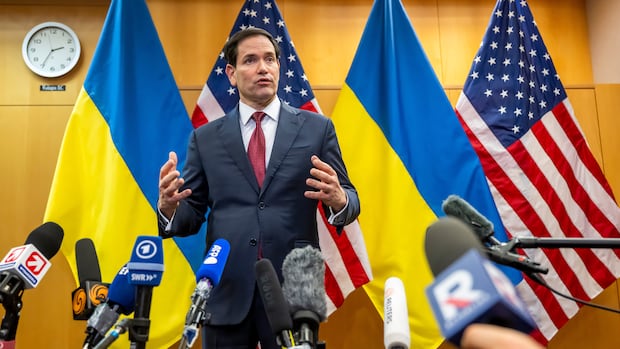 A man in a suit speaks in front of microphones with Ukrainian and American flags behind him.