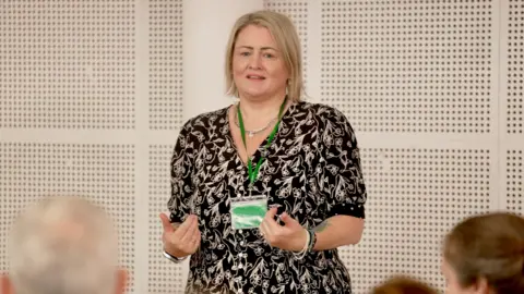 BBC/Stephen Fildes Fiona Wells, a woman with a blonde bob, wearing a black dress with a gold pattern, standing in front of an audience as she addresses a meeting of the adoptive parents group she founded.