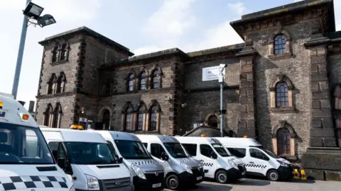 NIKLAS HALLE'N/AFP via Getty Images A general view of a Serco vehicle at Wandsworth prison in London. It is an imposing building with a gatehouse and what looks like a portcullis. A row of prison vans are by the entrance