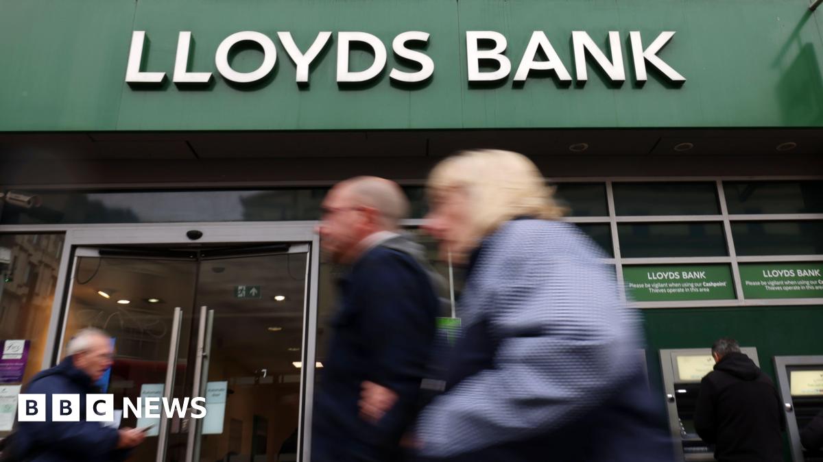 A shot of the front of a Lloyds Bank - it's branded name is in white lettering above the bank on its distinctive green background - in the foreground are a number of people walking and are out of focus, which draws focus to the bank's name - a person can also be seen taking money out of a cash machine