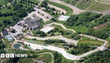 A birds eye view looking down on a dry ski slope snaking through a grass area with trees and buildings