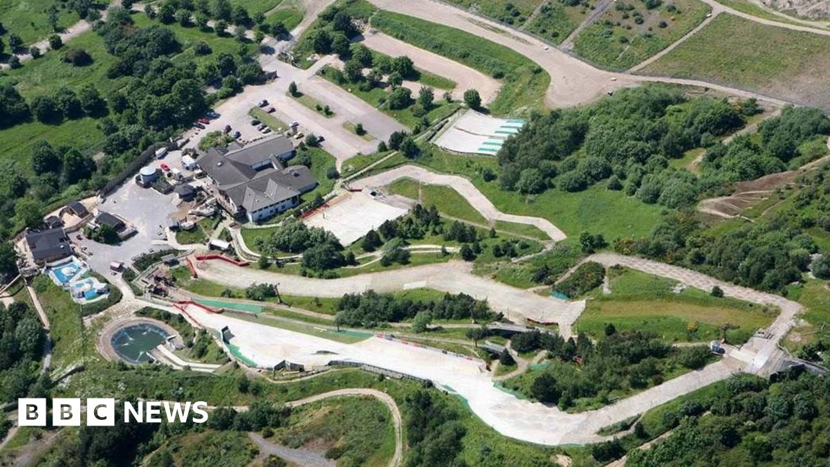 A birds eye view looking down on a dry ski slope snaking through a grass area with trees and buildings