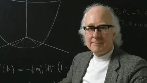 Open Prof Higgs is in front of a black board with calculations on it. He has white shoulder-length hair and black rimmed glasses. He is wearing a tweed suit jacket with a white polo-neck jumper underneath.