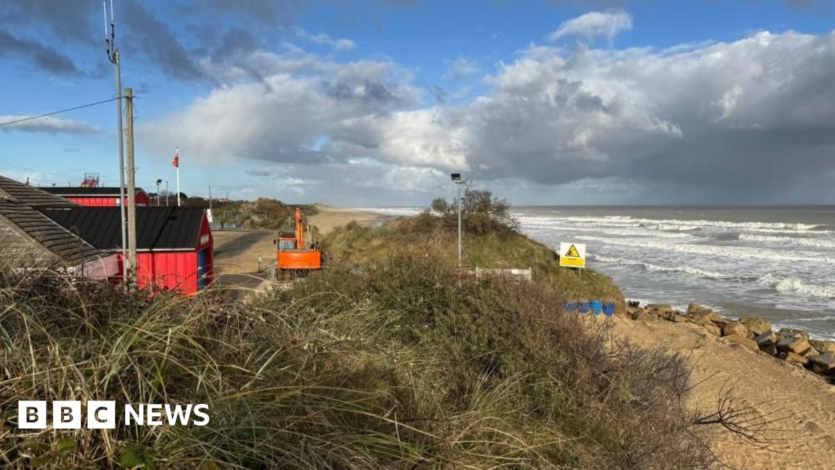 Hemsby beach and lifeboat station, with a digger on the beach repositioning concrete sea defence blocks. The lifeboat station is set back from the beach. It is a red building. The sea is at high tide, with white surf on the water.