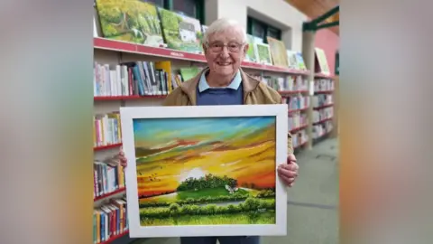 Robert Rose Robert Rose with library shelves behind him, holding a frames painting of a countryside sunset