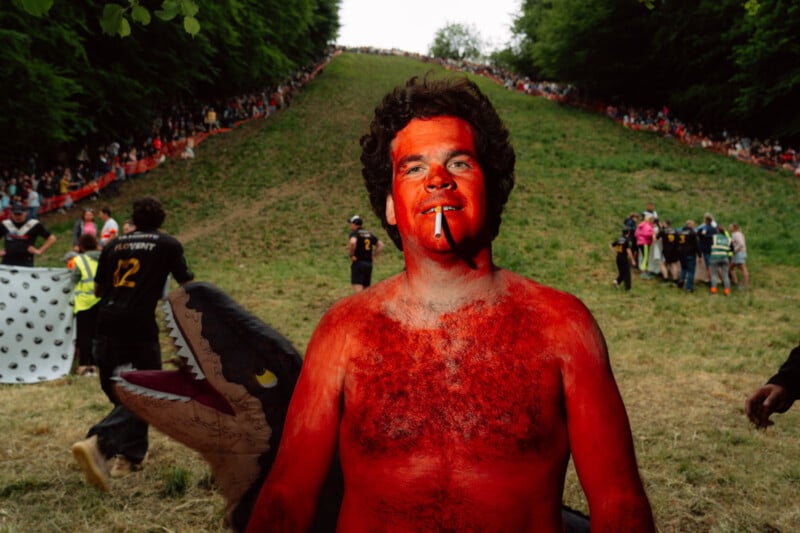 A shirtless man painted red with a cigarette in his mouth stands on a grassy slope during a crowded outdoor event, with people and a person in a shark costume visible in the background.