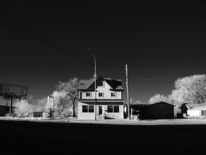 A two-story house with an attic stands alone under a dark sky, illuminated by bright sunlight. Leafless trees and power lines surround the building, and empty roads and other small structures are visible nearby.