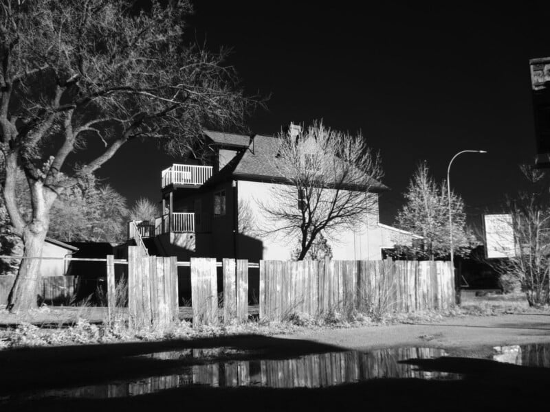 A black and white photo of a house with a porch and balcony, surrounded by leafless trees and a wooden fence. Shadows and puddles are visible in the foreground, reflecting part of the scene.