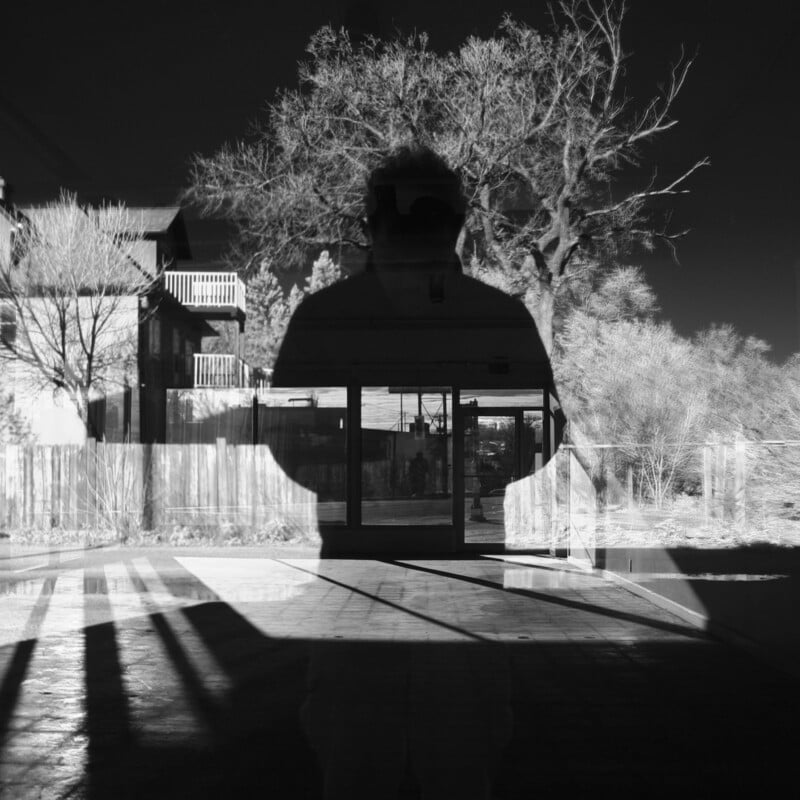 A black-and-white photo shows the silhouette of a person reflected in glass, blending with an outdoor scene of leafless trees, a building, and shadows on the ground, creating a layered, abstract effect.