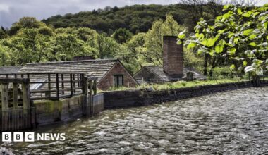 A dam with a stone wall in the foreground. Roofs of stone buildings can be seen beyond the wall.