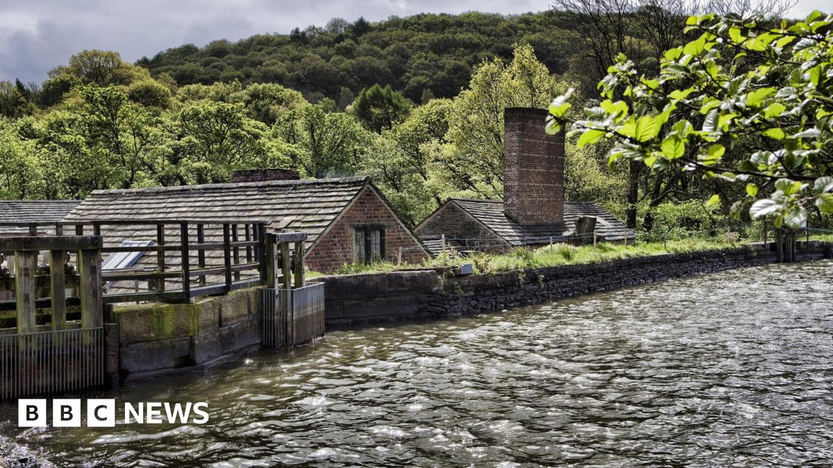 A dam with a stone wall in the foreground. Roofs of stone buildings can be seen beyond the wall.