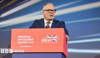 A man with short grey hair wearing a suit and thick black glasses, stood behind a red lectern with a red, white and blue Union Jack flag on it reading "Regional Investment Summit 2025 West Midlands".