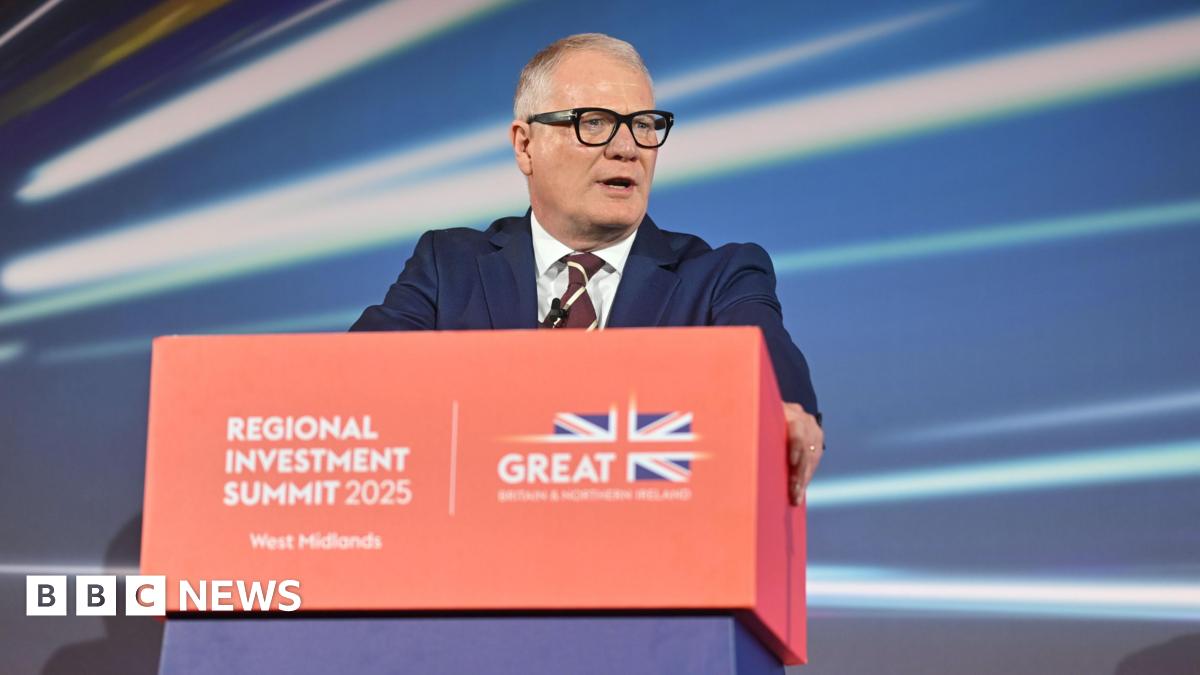 A man with short grey hair wearing a suit and thick black glasses, stood behind a red lectern with a red, white and blue Union Jack flag on it reading "Regional Investment Summit 2025 West Midlands".