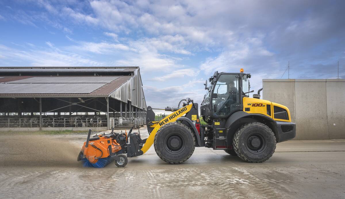 A promotional image of New Holland’s new W100D wheel loader using a broom attachment to sweep a large concrete apron with a dairy barn and cattle visible in the background.