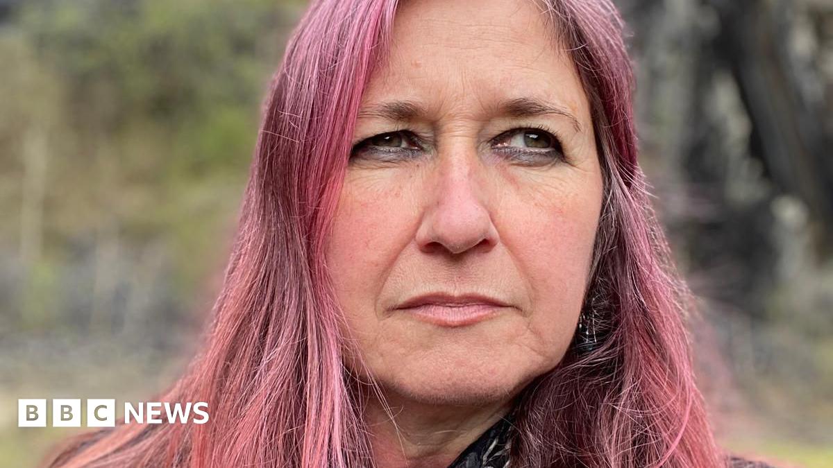 Helen looks into the distance, away from the camera, with a serious expression. She has long dyed pink hair, and is standing outside with blurred greenery behind her.