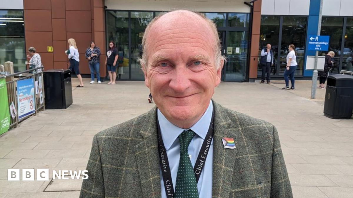 Nick Hulme is standing in front of Colchester hospital  in green checked jacket with rainbow pin and “Chief Executive” lanyard. He is smiling at the camera.