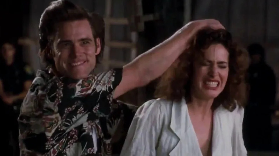 A scene from a movie featuring a man playfully pulling a woman's hair. The man wears a patterned shirt, the woman a light-colored suit