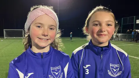 Two young girls are smiling for the camera. They are both wearing purple zip up jumpers with their football club branding on them in white. They are standing on a football pitch which is lit up with flood lights. 