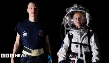 A man and women are stood next to each other. One is wearing a white space suit with a glass helmet, the other is wearing a navy blue T-shirt and has piping in his hand. They are both looking directly at the camera and stood in front of a black background.