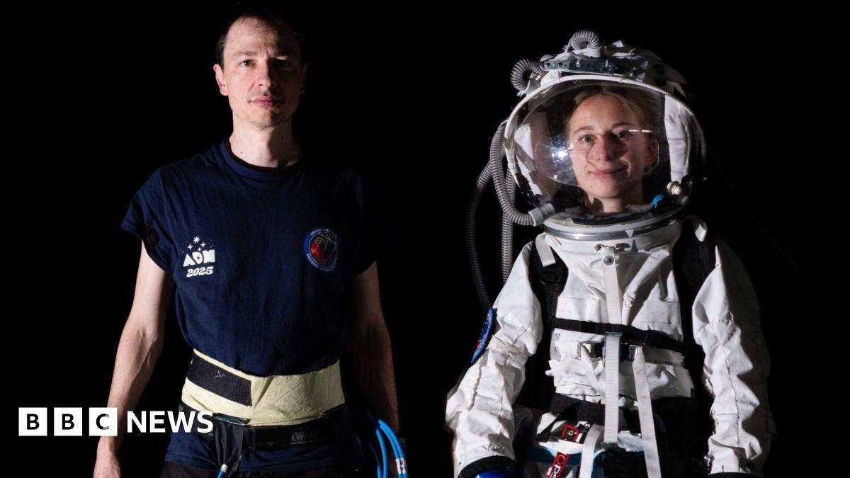 A man and women are stood next to each other. One is wearing a white space suit with a glass helmet, the other is wearing a navy blue T-shirt and has piping in his hand. They are both looking directly at the camera and stood in front of a black background.