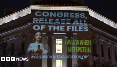 Wording is projected at nighttime onto a building that includes the phrase "Congress, release all files" in capital letters and a white font above a photo of a woman with long brown hair. The words "I was 14 when I met Epstein" are also seen in all capital letters and a green font