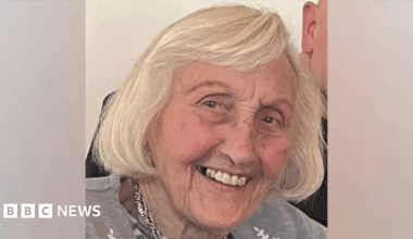 A family photo of Elizabeth. She has brown eyes  and shoulder length white hair. She is smiling at the camera. She is wearing a grey cardigan and a silver necklace.