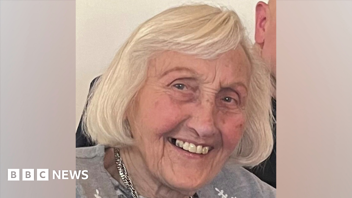 A family photo of Elizabeth. She has brown eyes  and shoulder length white hair. She is smiling at the camera. She is wearing a grey cardigan and a silver necklace.