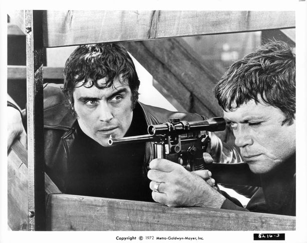 Ian McShane watching fellow acting hell raiser Oliver Reed aiming his weapon in a scene from the film 'Sitting Target', 1972