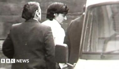 A black and white still from TV footage showing Peter Sullivan, who has thick black hair and is wearing a white shirt, being led to a police van in handcuffs.