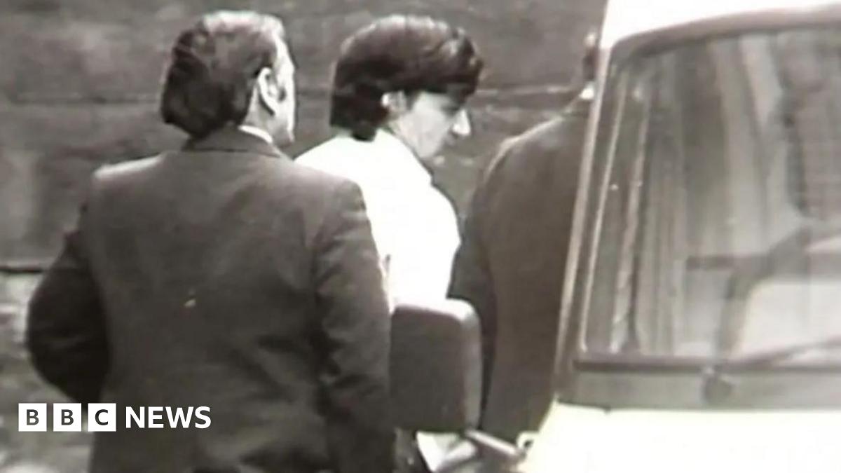 A black and white still from TV footage showing Peter Sullivan, who has thick black hair and is wearing a white shirt, being led to a police van in handcuffs.