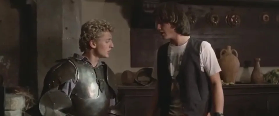 Two young men talking; one wears medieval armor, the other a casual vest and t-shirt. Scene suggests a mix of historical and modern attire